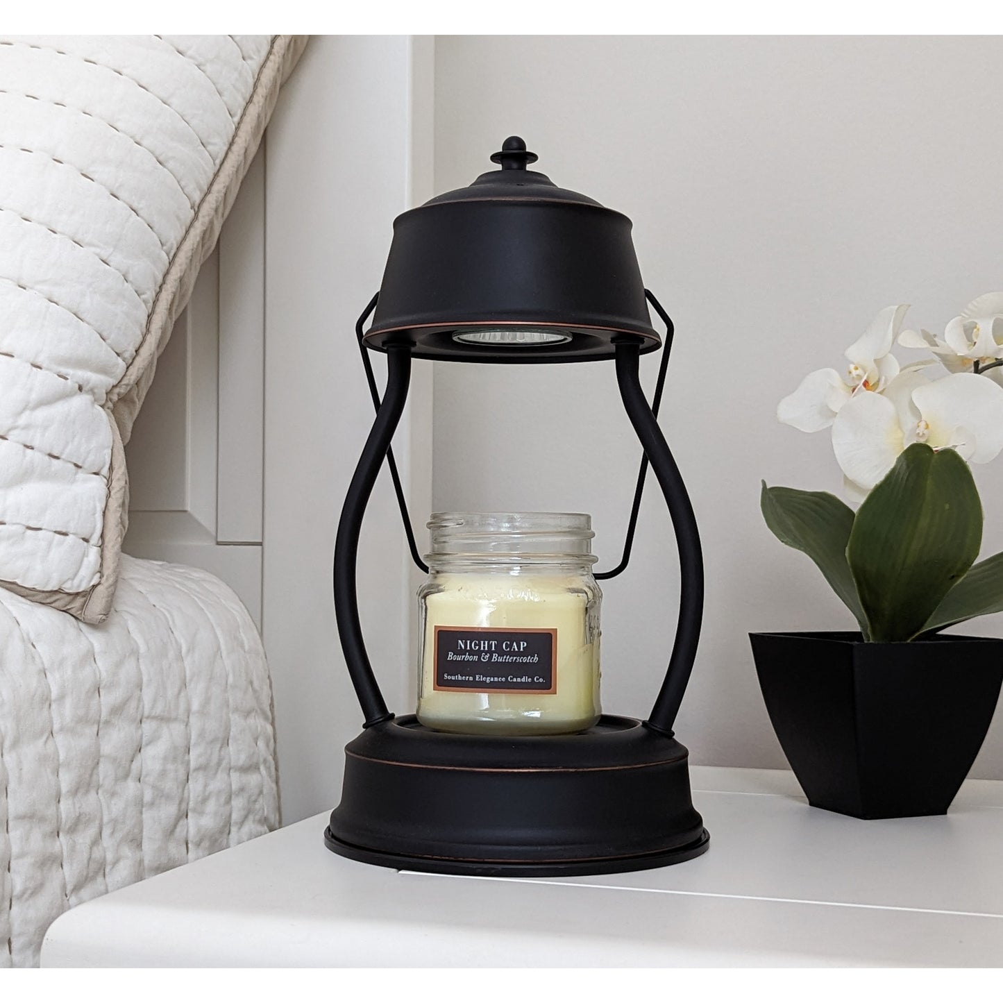 Image, unlit rubbed bronze candle warmer on a bedside table with small scented candle.
