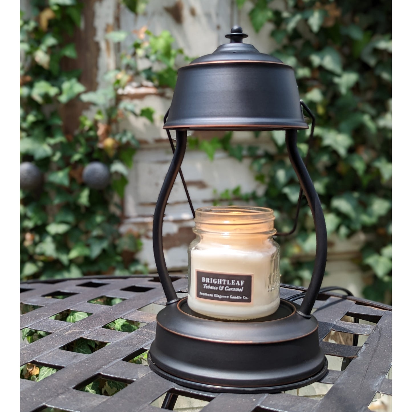 Image, rubbed bronze candle warmer on outdoor wrought iron table with lit small scented candle.